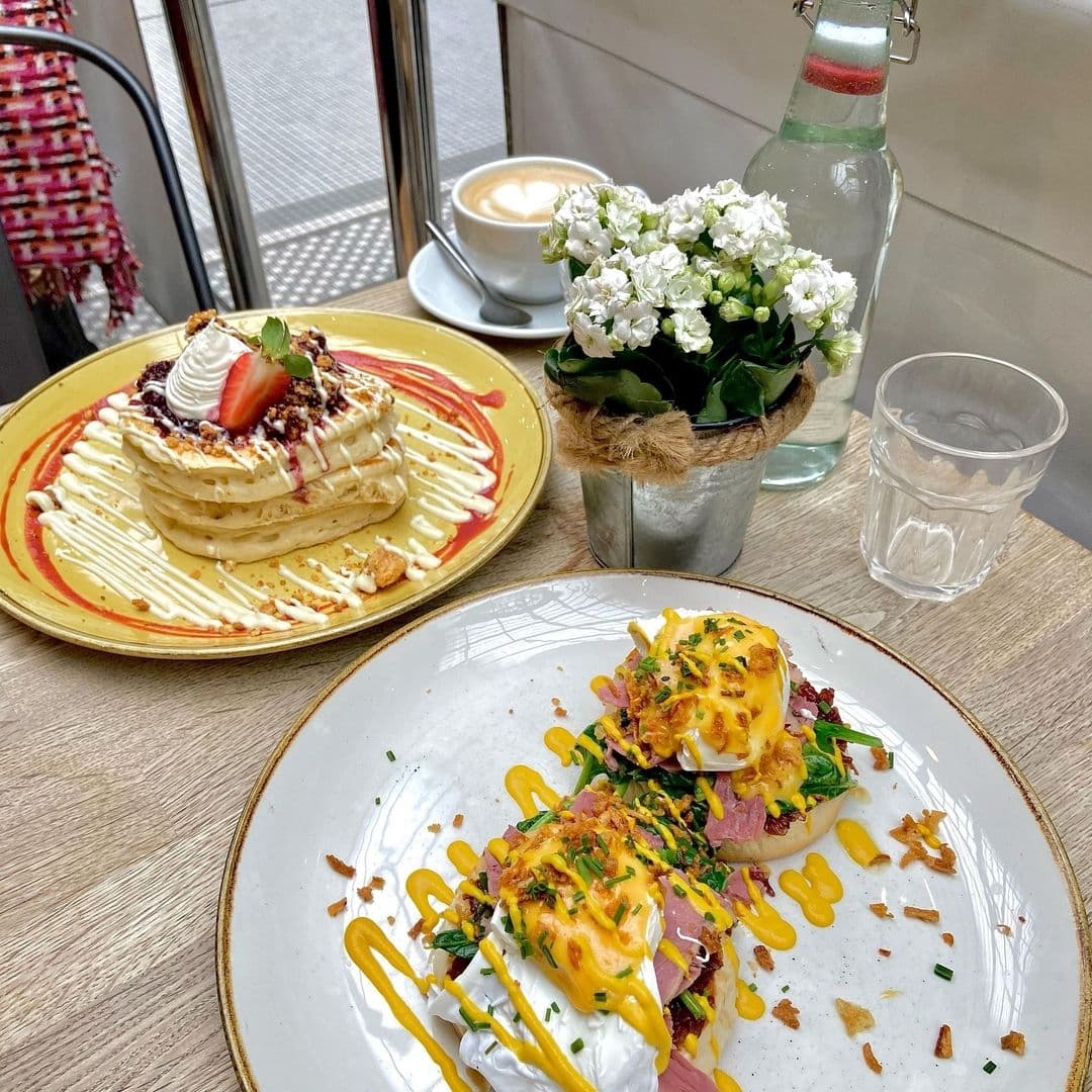 Olive Rye Brunch Pancakes Eggs Benedict Floral Decor Delicious breakfast spread at Olive & Rye featuring pancakes with whipped cream and strawberries, eggs benedict on English muffins, a cappuccino, and a small vase with white flowers.