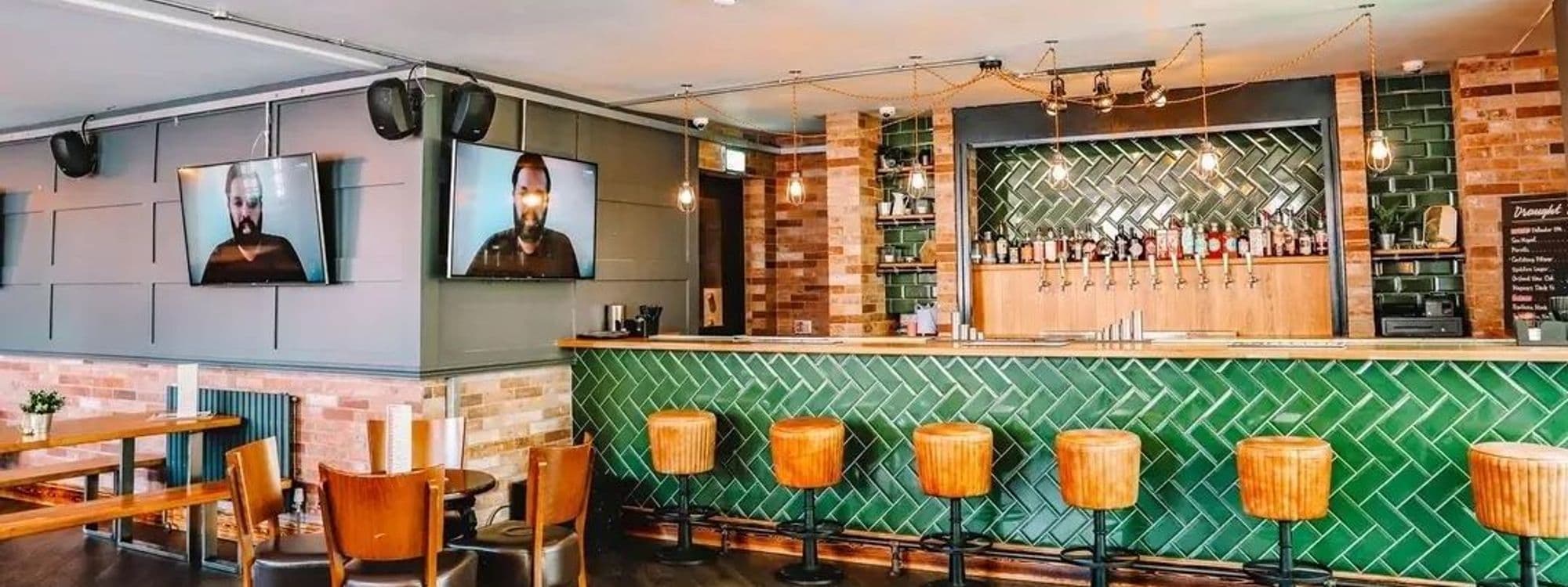 Interior of Parkside Tavern with a stylish bar area featuring green tiled walls, wooden bar stools, and a well-stocked bar with various drinks on tap. Wooden tables and chairs are arranged for guests, and two flat-screen TVs are mounted on the wall.