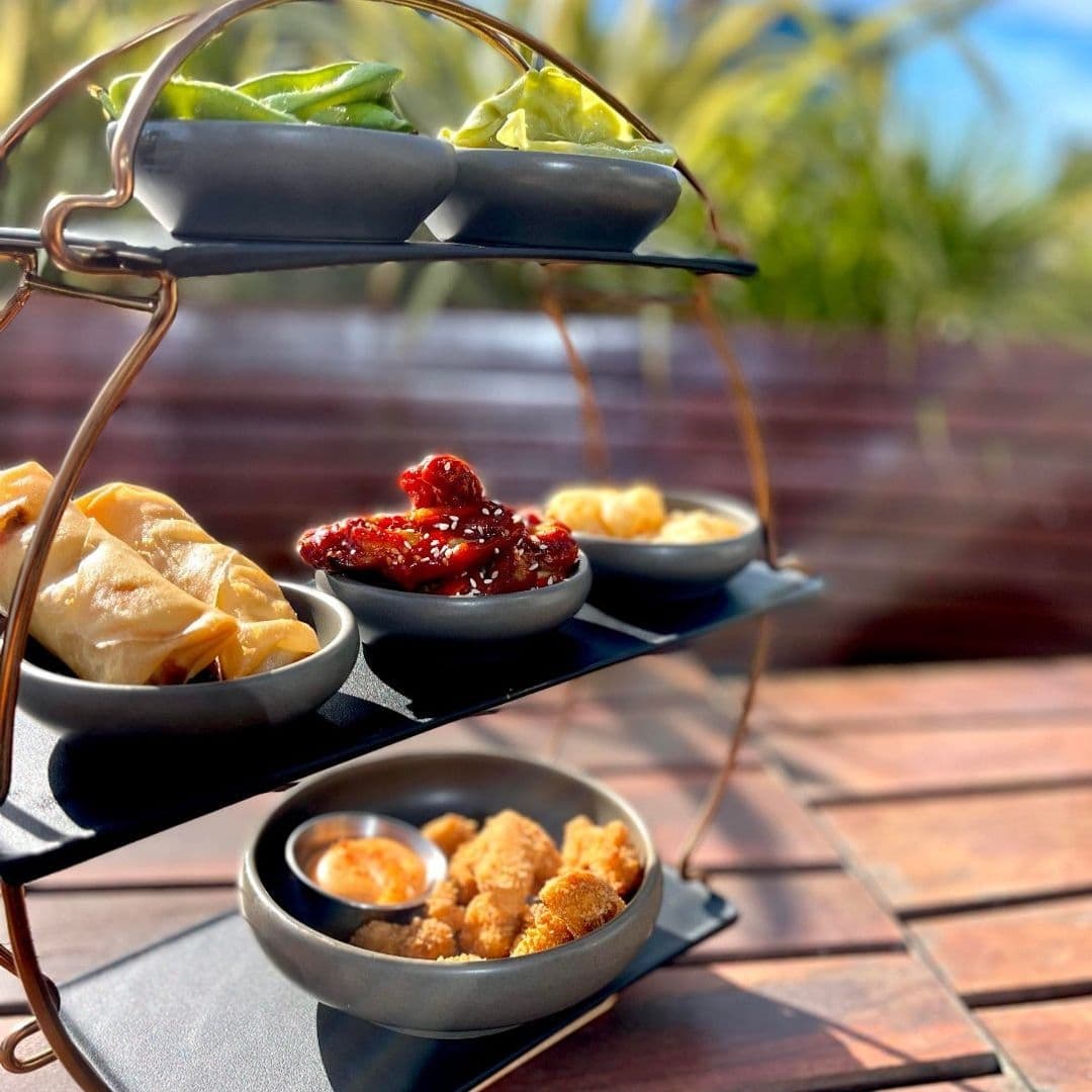 The Alchemist Trinity Gourmet Appetizers Outdoor Dining Three-tiered serving platter with appetizers at The Alchemist Trinity, featuring dumplings, glazed chicken wings, and breaded shrimp with dipping sauces, set on a wooden table with blurred greenery in the background.