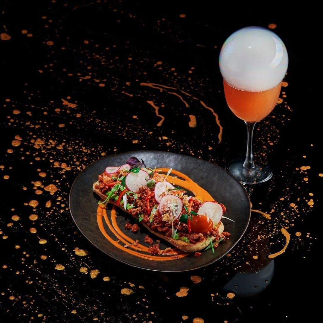 The Alchemist Trinity_Gourmet Dish Cocktail Presentation Gourmet dish at The Alchemist Trinity featuring an artistic presentation of sliced meats, vibrant vegetables, and puree on a black plate, accompanied by a cocktail with a spherical bubble garnish.
