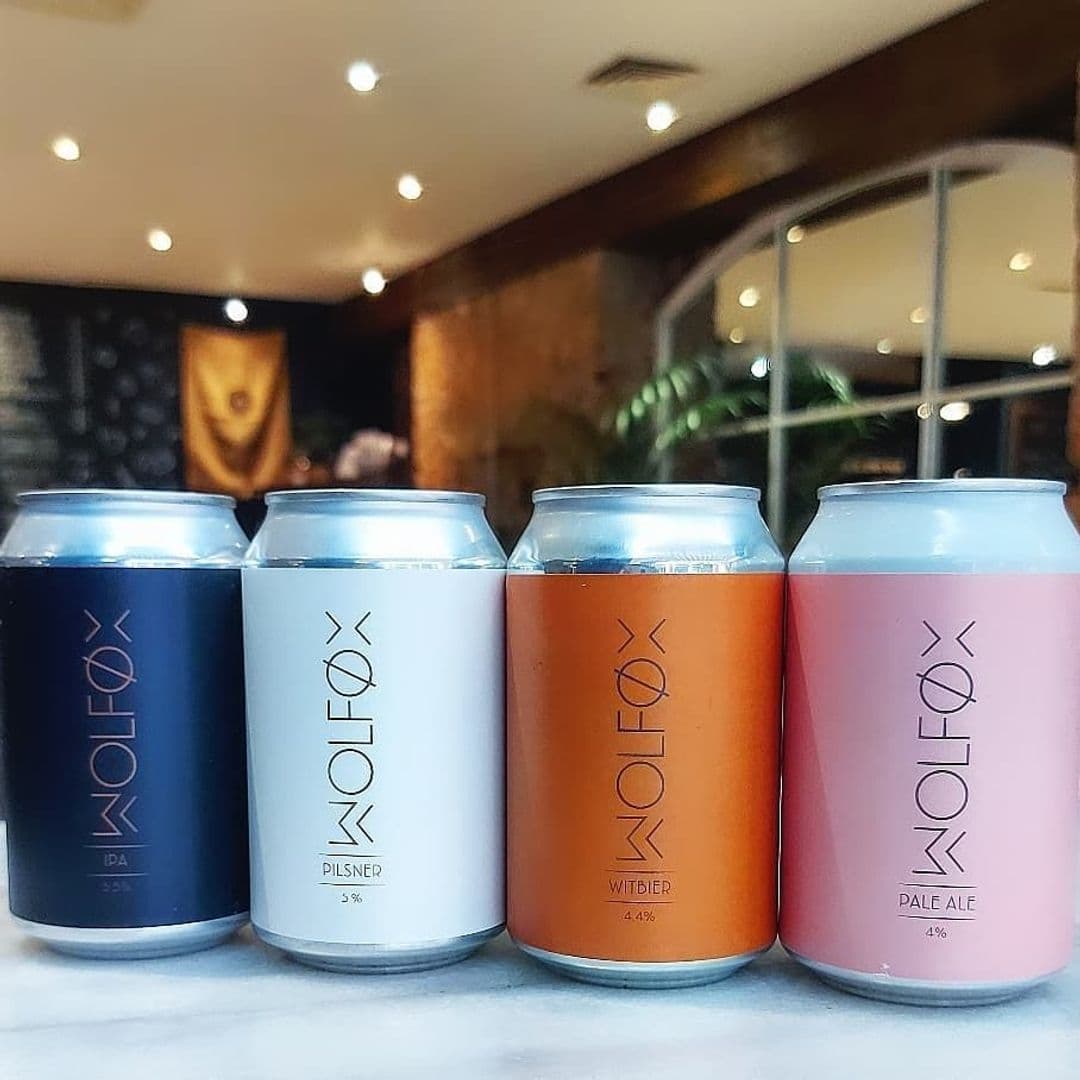 Wolfox_Coffee_ZAVIYA_Coffee_Bar_Restaurant_Craft_Beer_Cans_Display Four Wolfox craft beer cans on a table at ZAVIYA COFFEE Bar & Restaurant, with a blurred background featuring interior decor. The cans are labeled with different beer types: IPA, Pilsner, Witbier, and Pale Ale.