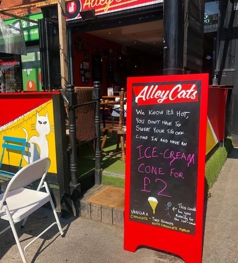 Alley Cats Cafe Bar Music Emporium Sunny Patio Ice Cream Sign Exterior view of Alley Cats Cafe Bar & Music Emporium with a red sidewalk sign advertising ice-cream cones for £2, featuring a cartoon cat, against a sunny backdrop with outdoor seating.