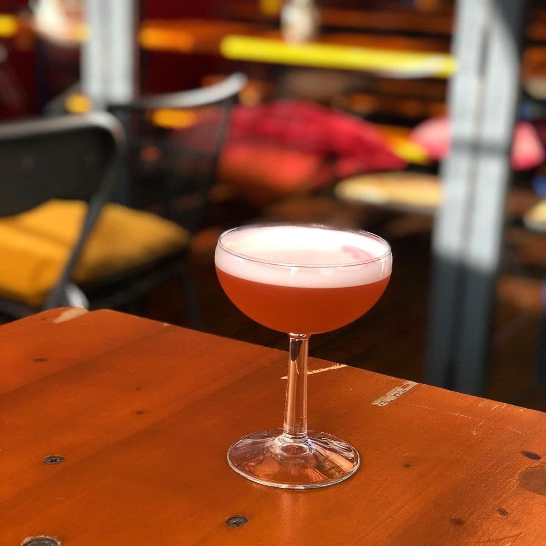 Alley Cats Cafe Bar Music Emporium_Cocktail Glass Bokeh Background Pink cocktail in a coupe glass on a wooden table at Alley Cats Cafe Bar & Music Emporium, with blurred background of colorful chairs.