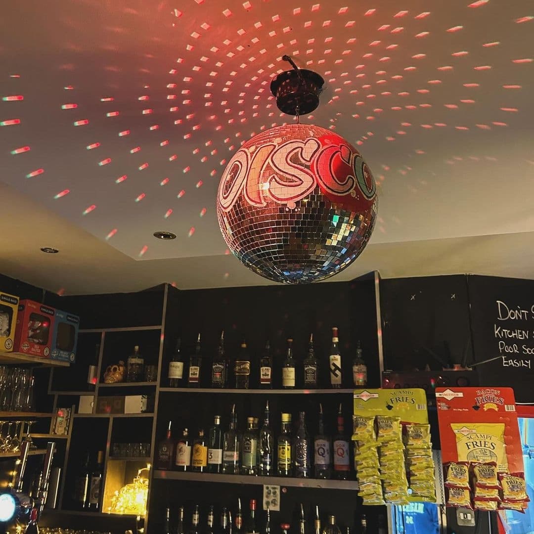 Alley Cats Cafe Bar & Music Emporium_Disco_Ball_Bar_Shelves Interior of Alley Cats Cafe Bar & Music Emporium featuring a large disco ball with the word "DISCO" reflecting red and white lights on the ceiling, behind a bar stocked with various bottles of alcohol and snacks.