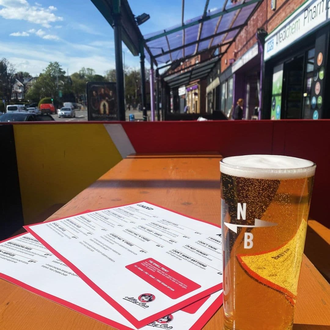 Alley Cats Cafe Bar Music Emporium_Outdoor Seating Sunny Day Cold Beer Outdoor seating at Alley Cats Cafe Bar & Music Emporium with a close-up of a pint of beer and a menu on a wooden table, overlooking a sunny street scene with shops and pedestrians.