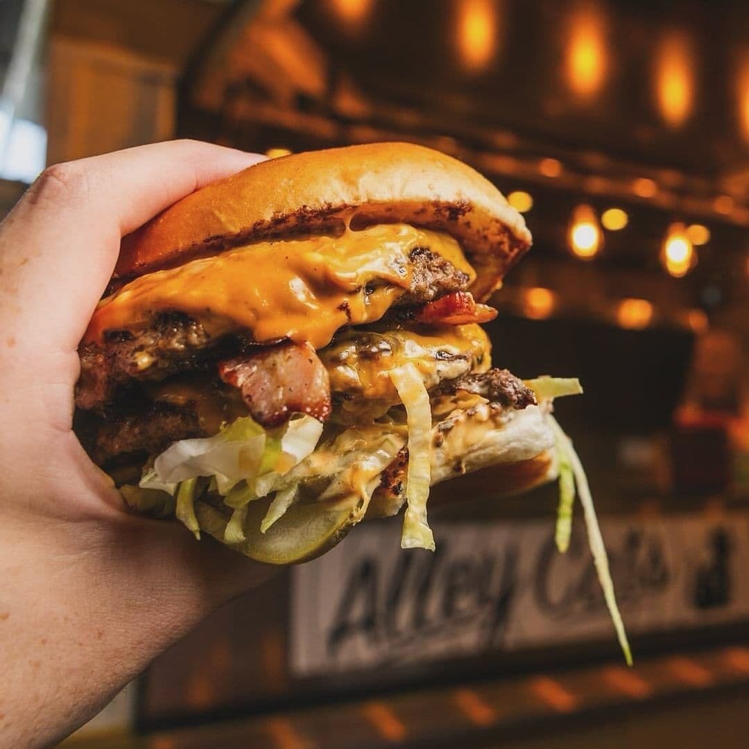 Alley_Cats_Cafe_Bar_Music_Emporium_Juicy_Bacon_Cheeseburger Juicy double cheeseburger with bacon, lettuce, and sauce held in a hand at Alley Cats Cafe Bar & Music Emporium, with warm ambient lighting in the background.