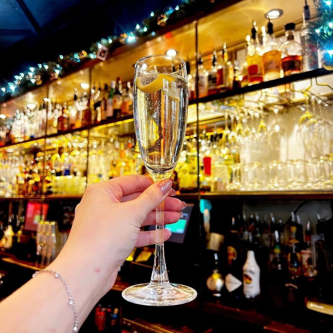 Be At One Boar Lane Champagne Celebration Bar Atmosphere Hand holding a champagne flute with a slice of lemon at Be At One – Boar Lane, with a blurred background of a well-stocked bar and festive lights.