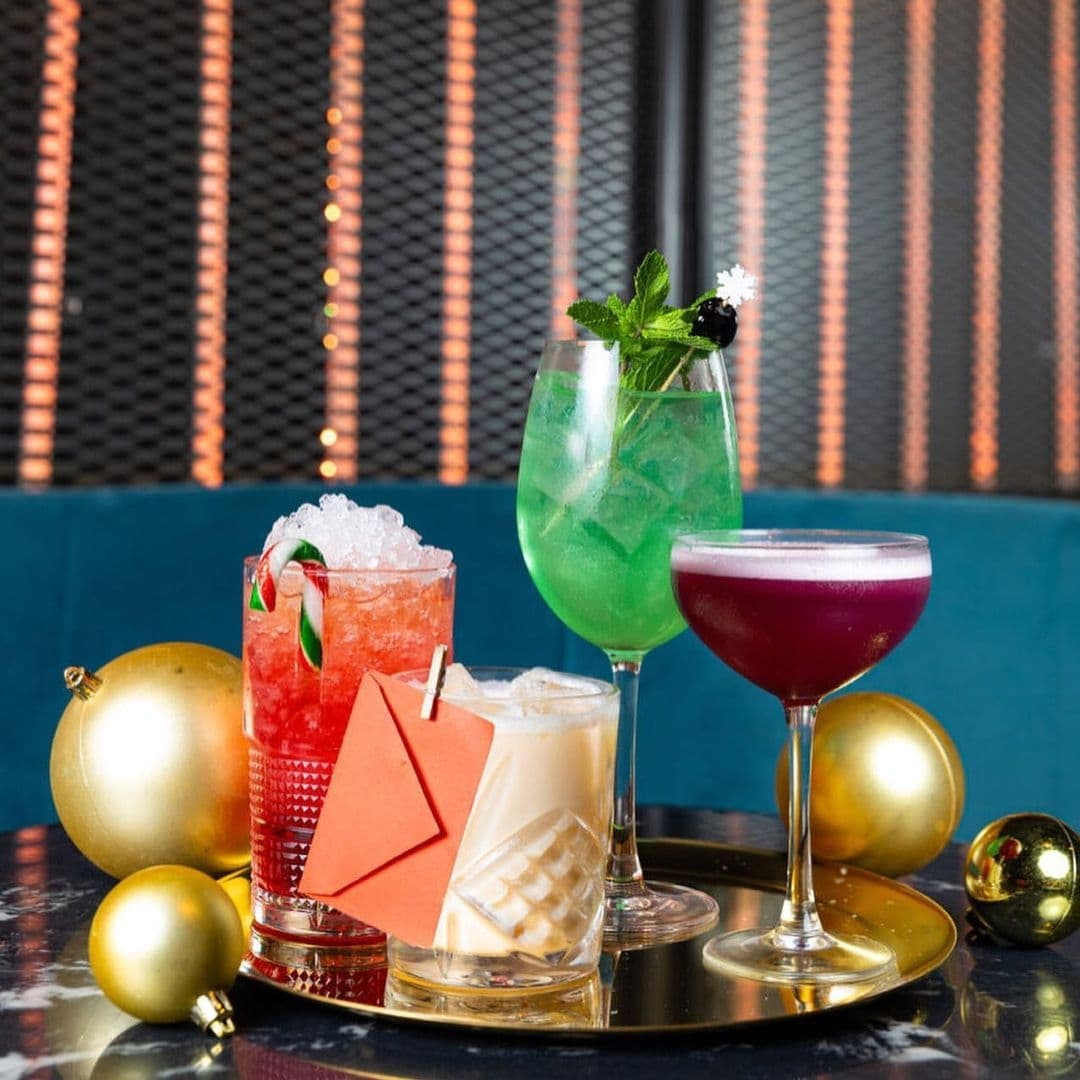 Be At One Boar Lane Cocktails Colorful Decor Festive Atmosphere Alt text: "Assorted colorful cocktails on a tray at Be At One – Boar Lane, with a festive decoration of golden baubles, set against a backdrop of a teal blue booth and warm, orange vertical light accents."