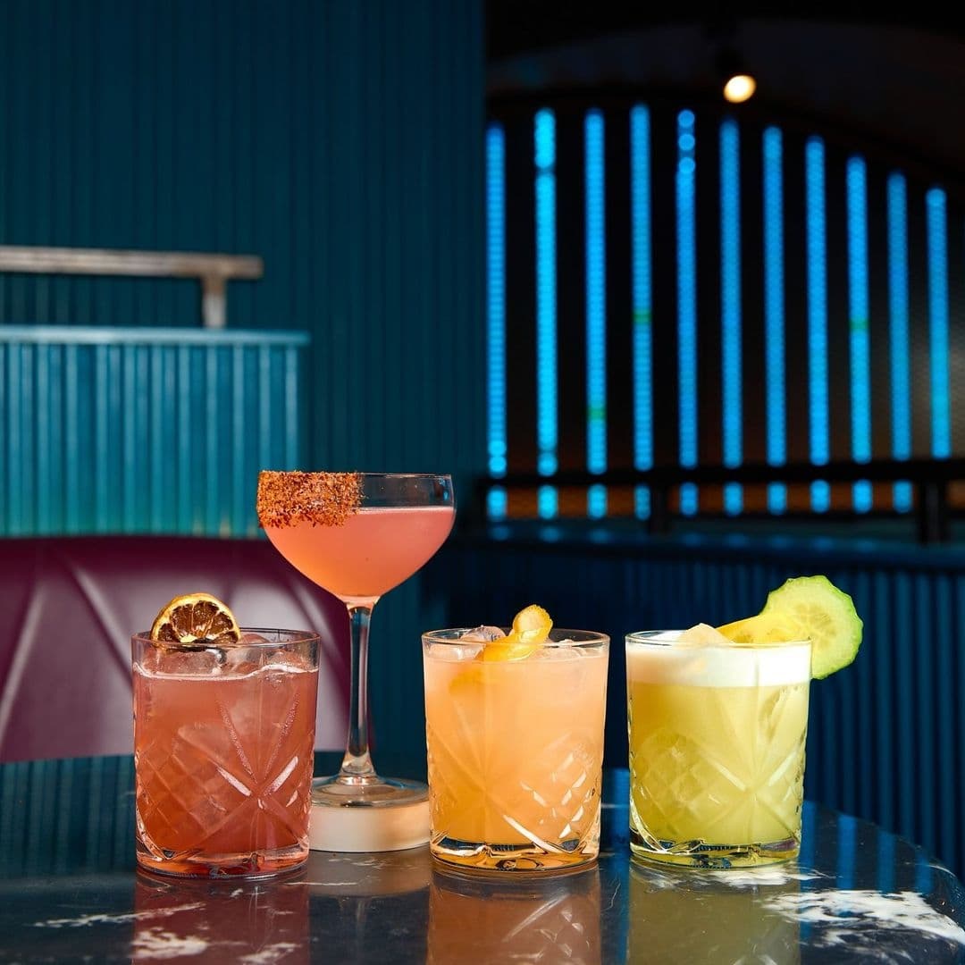 Be At One Boar Lane Cocktails Colorful Drinks Bar Interior Assorted colorful cocktails on a table at Be At One – Boar Lane with a teal blue backdrop and ambient lighting.