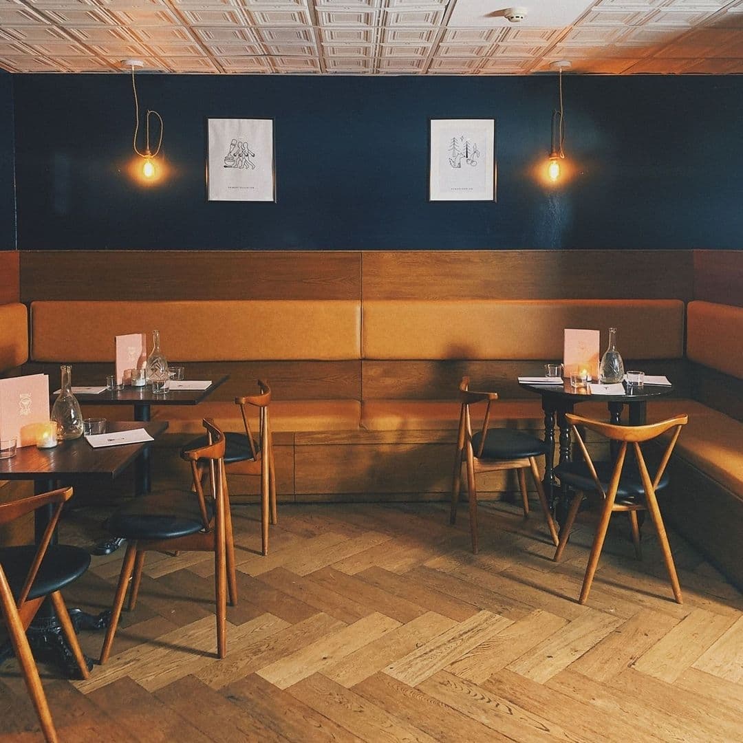 Below Stairs Cozy Restaurant Interior Wooden Furniture Modern Decor Interior of Below Stairs restaurant with wooden tables and chairs, a cozy booth with cushions, and framed artwork on a navy blue wall, illuminated by warm pendant lighting.