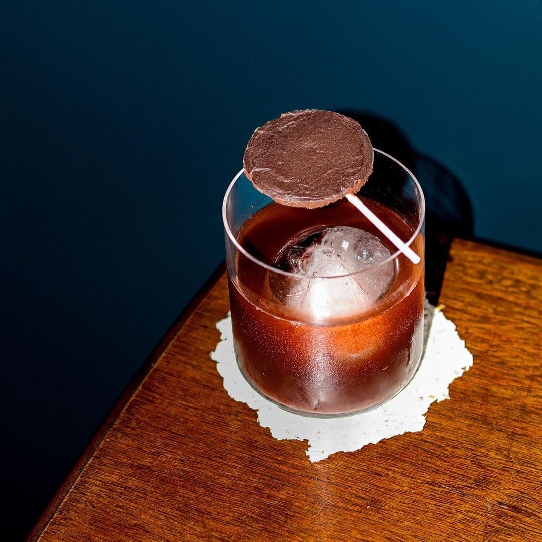 Below Stairs Signature Cocktail Chocolate Garnish Wooden Table Craft cocktail with a chocolate garnish at Below Stairs, presented on a wooden surface against a dark blue background.