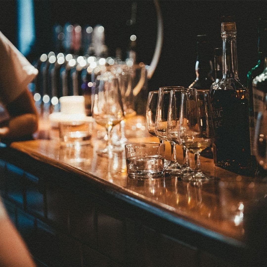 Below Stairs_Cozy Bar Atmosphere_Glasses Bottles Wooden Counter Close-up of a wooden bar counter at Below Stairs with a selection of glasses and bottles, suggesting a cozy and inviting atmosphere for drinks and socializing.