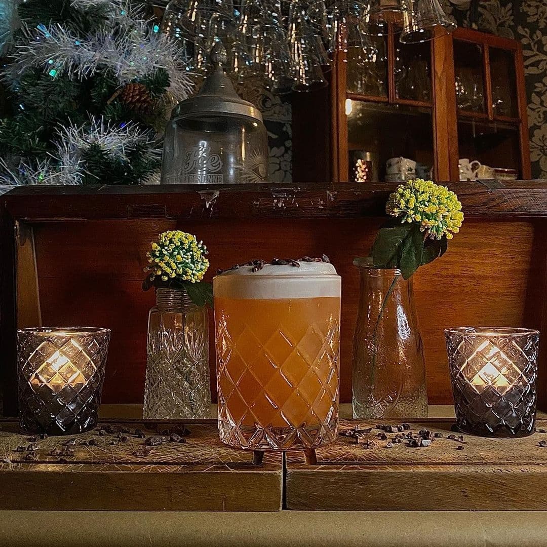 Blind Tyger Cocktail Candlelight Atmosphere Cozy ambiance at Blind Tyger with a festive cocktail in a diamond-patterned glass, flanked by lit candles and small yellow flowers, set against a backdrop of a Christmas tree and vintage decor.