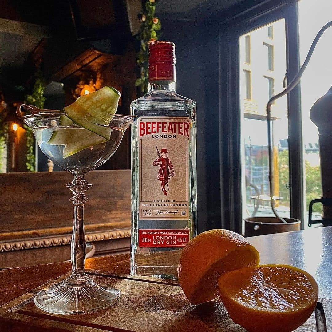 Blind Tyger Gin Cocktail Citrus Wooden Table Bottle of Beefeater London Dry Gin with cocktail glass garnished with cucumber slices on a wooden table at Blind Tyger, with a sliced orange in the foreground and a window in the background.