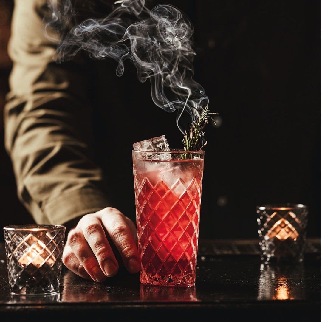 Blind Tyger Smoky Cocktail Ambiance A person's hand resting on a bar next to a smoky, red cocktail in a cut-glass tumbler garnished with a sprig of rosemary, with two votive candles in the background, at Blind Tyger.