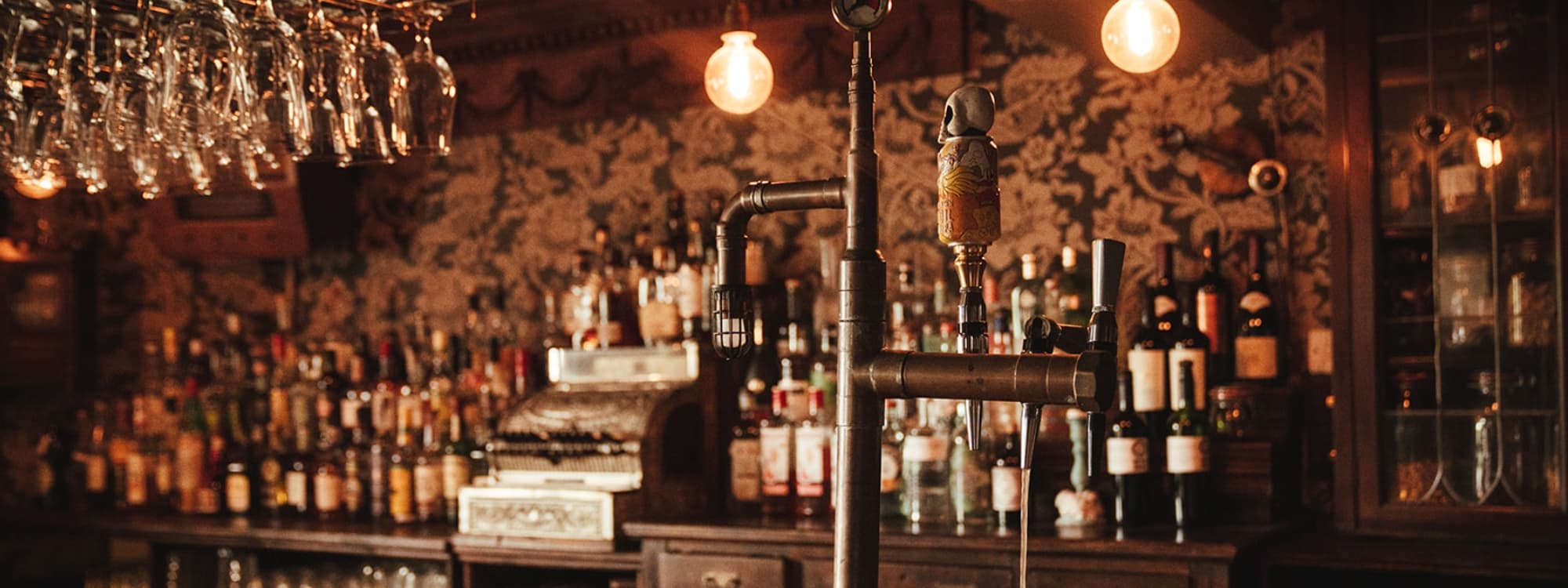 Interior of Blind Tyger showing a vintage-style bar with hanging glasses, a variety of liquor bottles, and warm ambient lighting.
