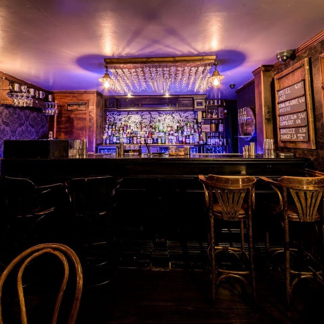 Blind Tyger Vintage Bar Interior Lighting Atmosphere Interior of Blind Tyger, featuring a dimly lit bar with an array of bottles, elegant glassware hanging above, and vintage-style wooden stools in the foreground.