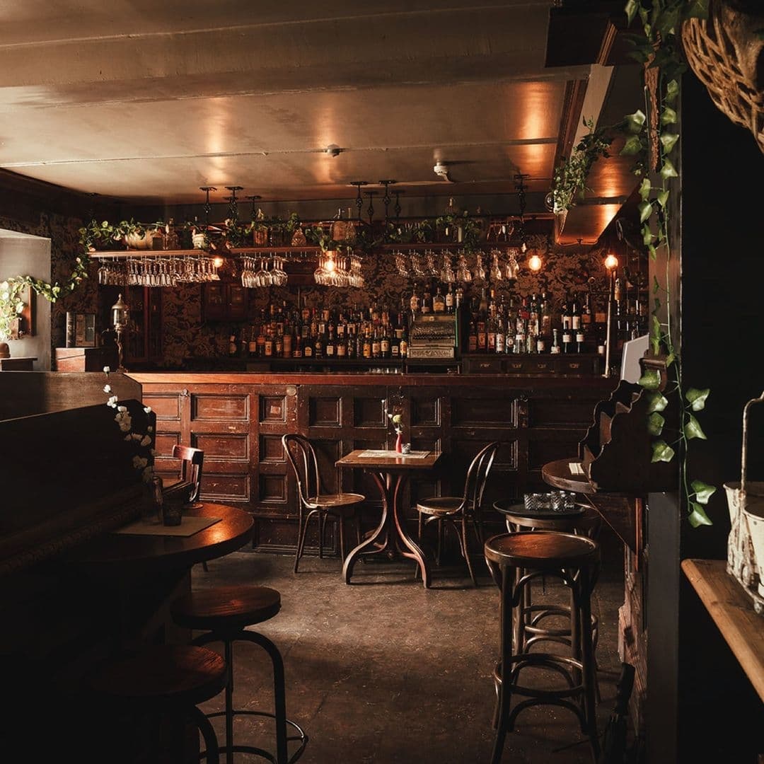Blind Tyger Vintage Speakeasy Atmosphere Bar Interior of Blind Tyger, showcasing a cozy bar area with wooden stools, tables, a well-stocked bar with an array of bottles, dim lighting, and decorative greenery.