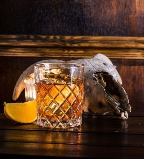 Blind Tyger Whiskey Glass Citrus Skull Backdrop Amber-colored cocktail in a cut-glass tumbler with a slice of orange, beside an animal skull, on a wooden surface at Blind Tyger.