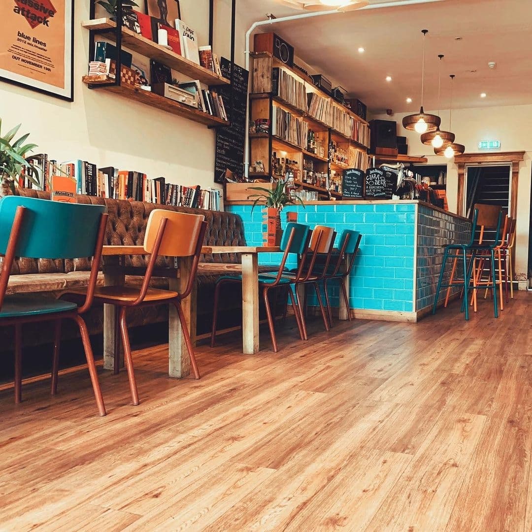 Melodie71_CozyCafeInterior_ColorfulChairs_WoodenFloor Interior of Melodie 71 featuring a cozy cafe setting with colorful chairs, a wooden bar counter with blue tiles, bookshelves, and pendant lighting.