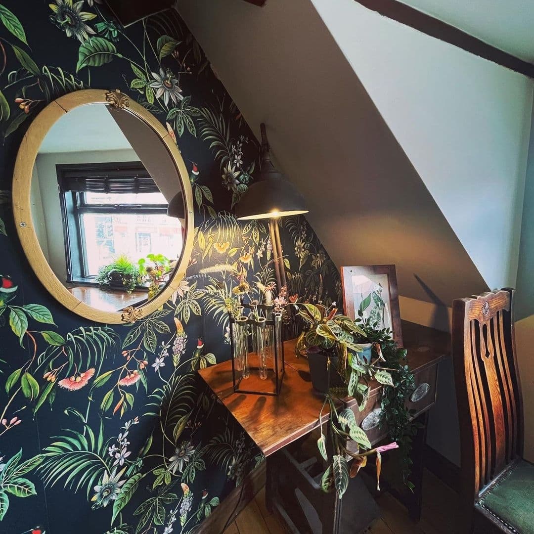 Number 8_Botanical Wallpaper Vintage Decor Desk Area Cozy corner at Number 8 featuring a botanical-themed wallpaper, a round mirror with a golden frame, a wooden desk with plants, and a vintage-style lamp.
