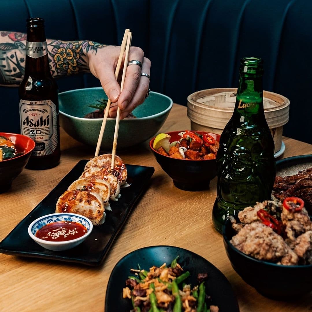 Power_Corruption_Lies_Asian_Cuisine_Dining_Experience Alt text: "Diverse Asian cuisine at Power, Corruption & Lies, featuring a person using chopsticks to eat gyoza, with dishes like spicy tofu, steamed dumplings, and fried meat, accompanied by bottles of Asahi beer and Lucky Buddha lager."
