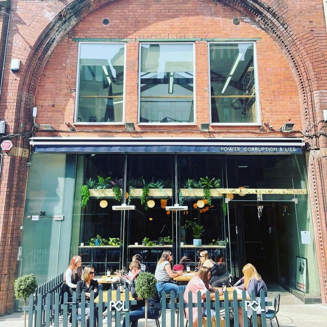 Power_Corruption_Lies_Outdoor_Dining_Brick_Architecture Exterior view of Power, Corruption & Lies restaurant with patrons dining at outdoor tables, lush plants decorating the facade, under clear skies.
