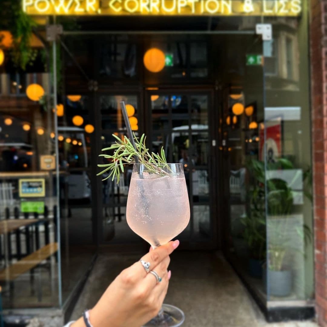 Power_Corruption_and_Lies_Cocktail_Handheld_Entrance A hand holding a stemmed glass with a pink beverage and a sprig of rosemary, with the entrance to "Power, Corruption & Lies" visible in the background.