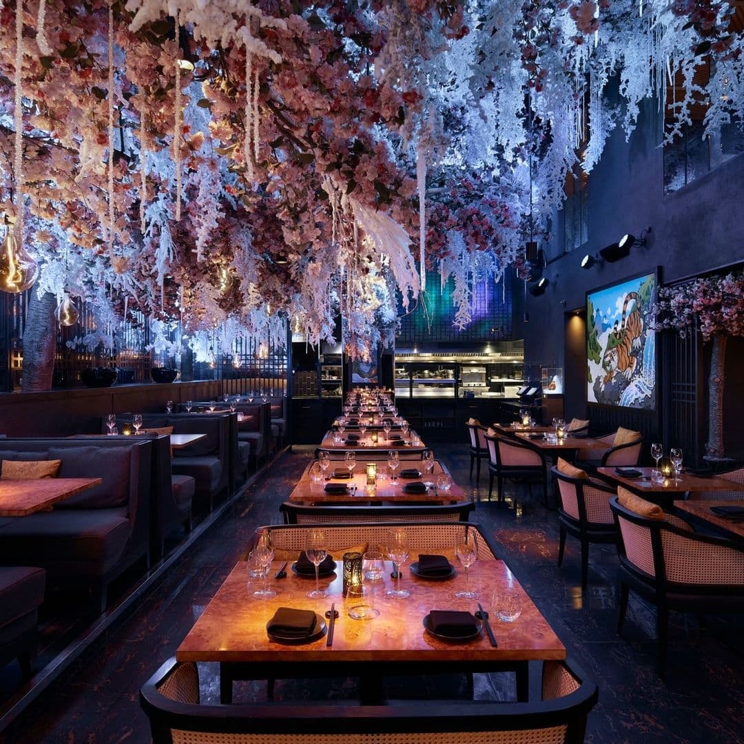 Tattu_Elegant_Interior_Cherry_Blossoms_Dining_Ambiance Elegant dining area at Tattu with tables set for service, adorned with cherry blossoms and icicle-like decorations hanging from the ceiling, ambient lighting, and a vibrant mural on the wall.