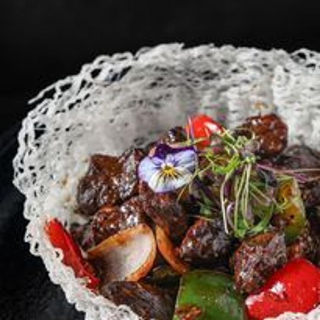 Tattu_Gourmet_Asian_Cuisine_Elegant_Presentation Gourmet dish at Tattu featuring glazed meat cubes with colorful bell peppers, garnished with a delicate edible flower and microgreens, presented on an intricate white lace-like doily over a dark background.