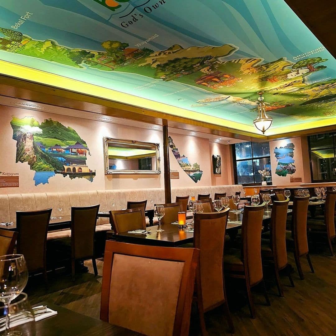 Tharavadu Restaurant Interior Elegant Dining Experience Interior of Tharavadu restaurant showing dining tables set with glasses and napkins, warm lighting, and walls decorated with colorful murals depicting landscapes.
