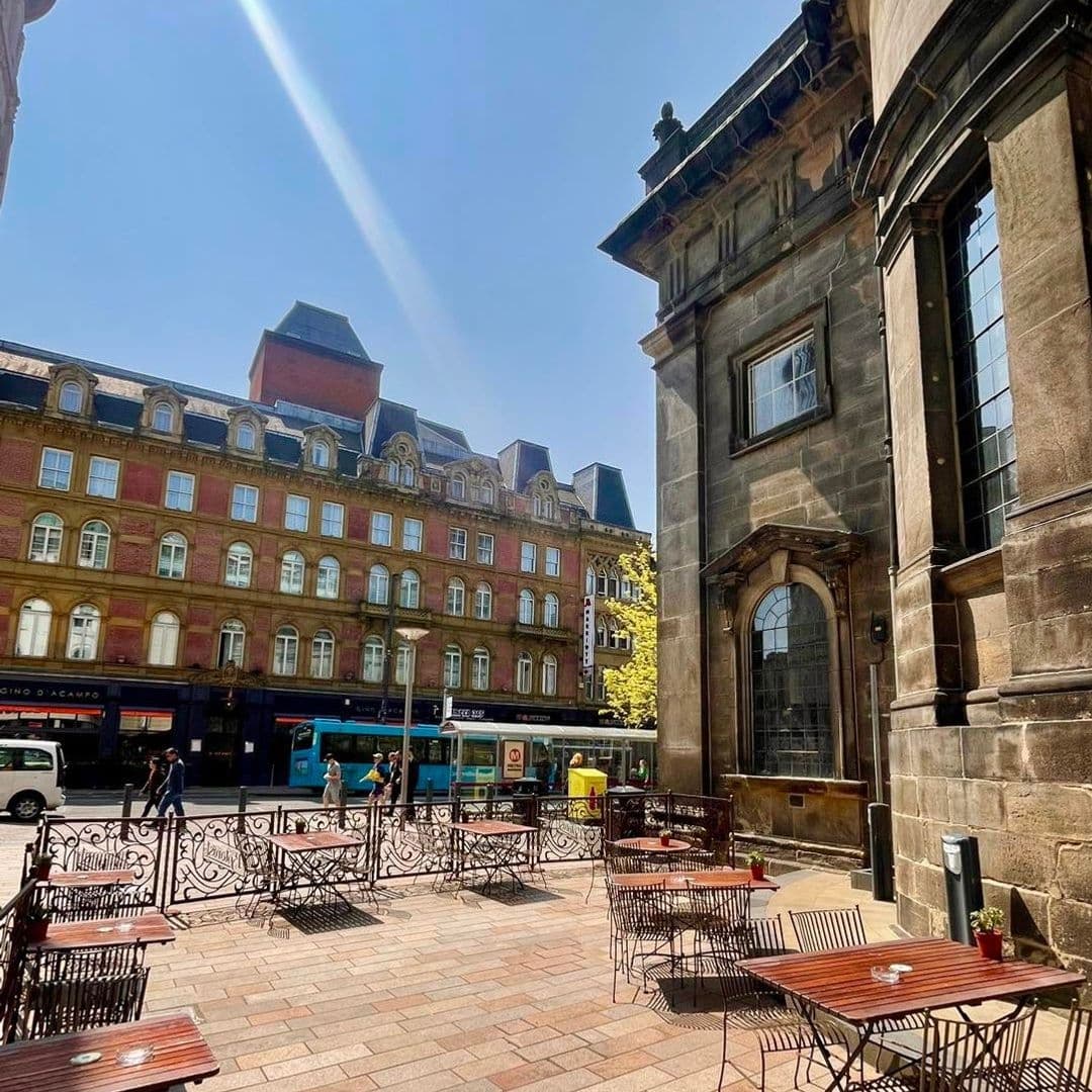 The Botanist Trinity Sunny Outdoor Seating Architecture Street View Outdoor seating area of The Botanist Trinity with wrought-iron furniture on a sunny day, historic buildings in the background, and a clear blue sky.