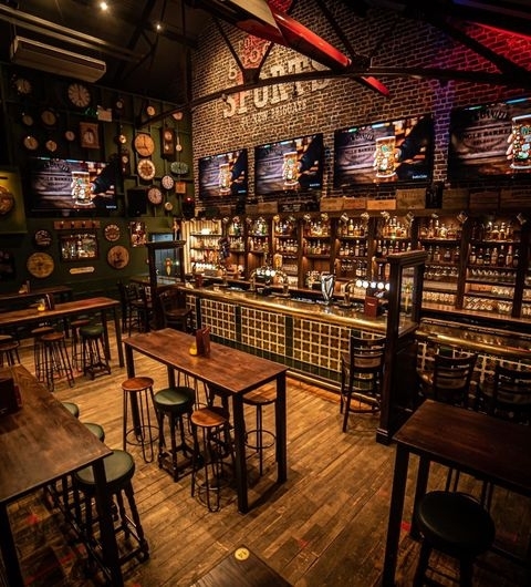 The Brotherhood of Pursuits and Pastimes Classic Pub Interior Wooden Furniture Warm Lighting Interior of The Brotherhood of Pursuits and Pastimes showing a well-stocked bar with multiple liquor bottles, wooden stools, high tables, and multiple television screens on a brick wall.