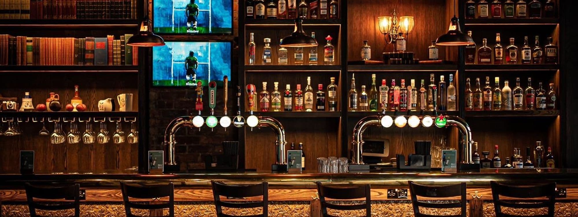 Interior view of The Brotherhood of Pursuits and Pastimes showing a well-stocked bar with multiple beer taps, shelves filled with a variety of spirits, and sports broadcasting on television screens, all set against a backdrop of dark wood and ambient lighting.
