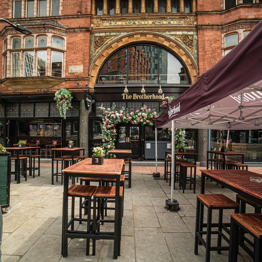 The Brotherhood of Pursuits and Pastimes Outdoor Seating Architecture Floral Decor Outdoor seating area of The Brotherhood of Pursuits and Pastimes with wooden tables and stools, floral decorations, and an ornate building facade in the background.