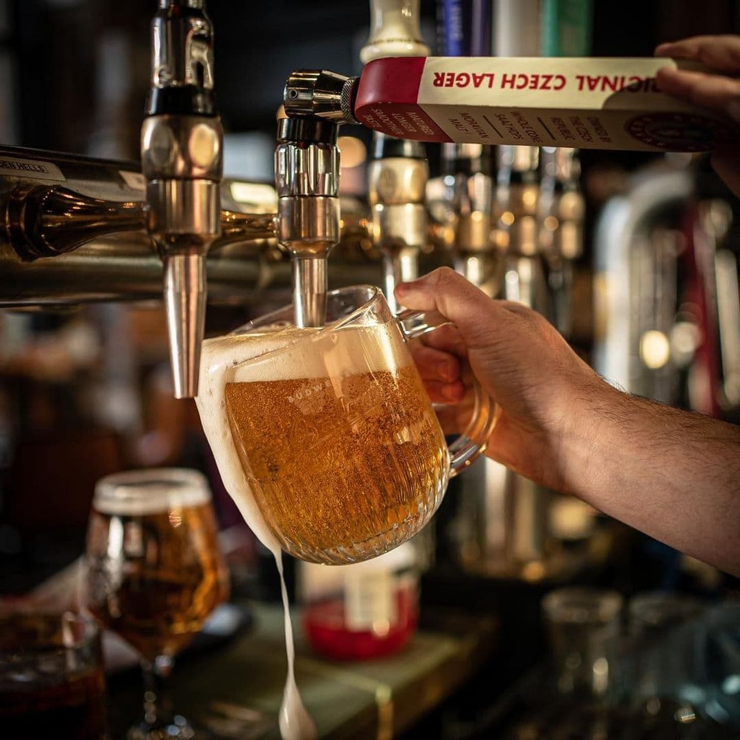 The Brotherhood of Pursuits and Pastimes_Beer Pouring Tap Frothy Glass A bartender pouring a golden beer from a tap into a clear glass mug at The Brotherhood of Pursuits and Pastimes.