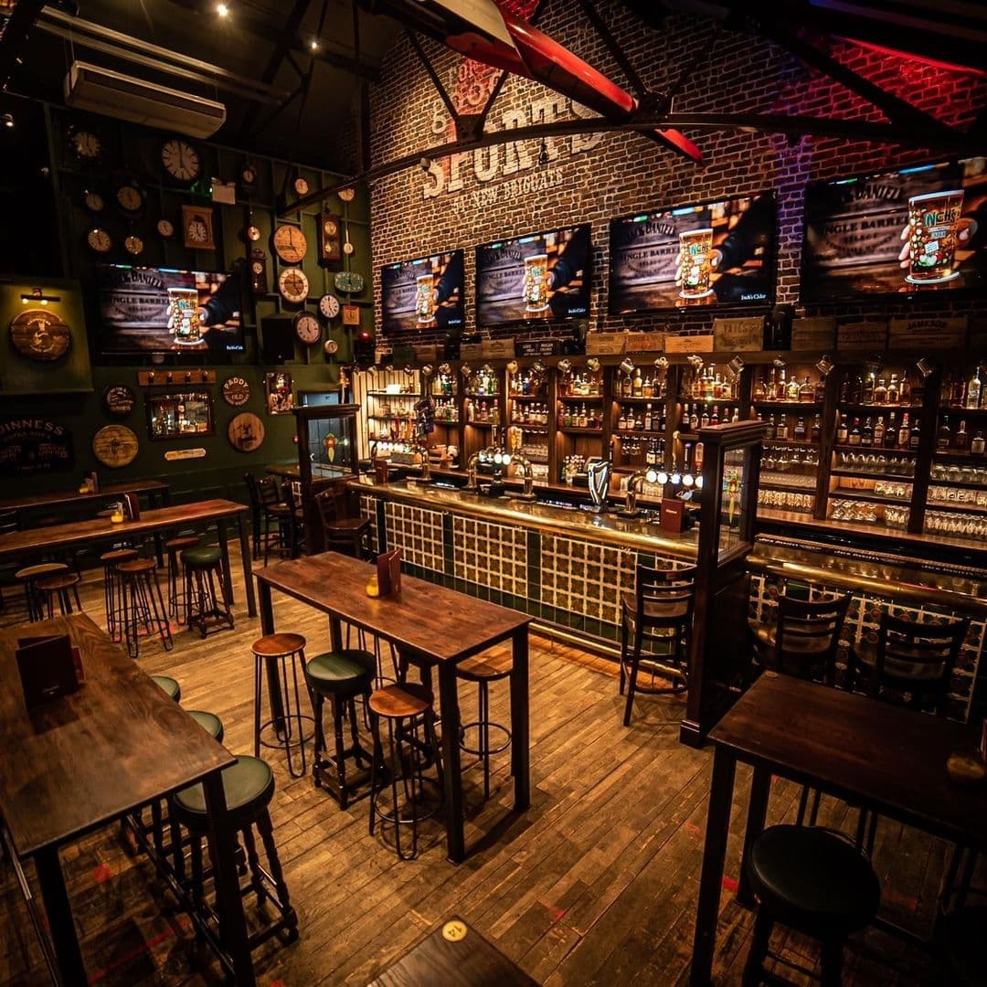 The Brotherhood of Pursuits and Pastimes_Cozy Pub Interior Wood Bar Brick Walls Interior of The Brotherhood of Pursuits and Pastimes showing a cozy bar area with wooden tables and stools, a well-stocked bar counter, multiple television screens displaying sports, and walls adorned with dartboards and vintage decor.