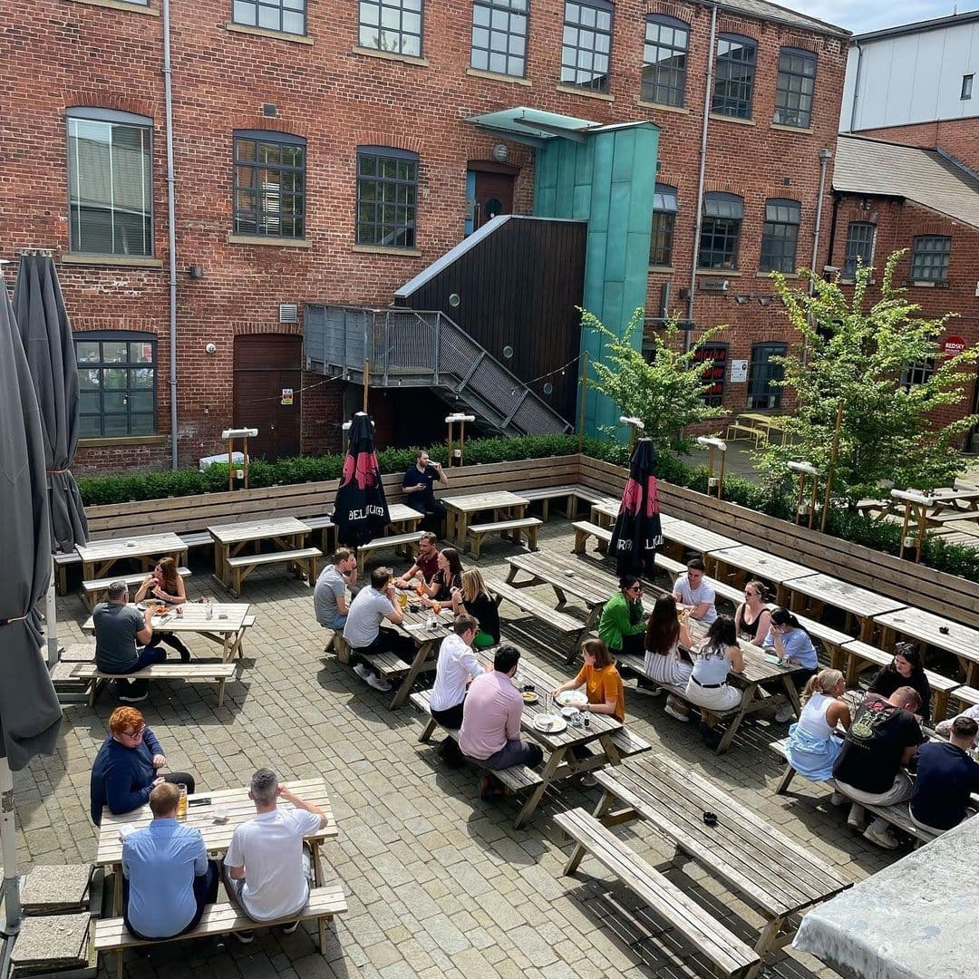 The Cross Keys Outdoor Patio Dining Socialising Brick Building Outdoor seating area at The Cross Keys with people socializing at wooden picnic tables on a sunny day, surrounded by red brick buildings with greenery.