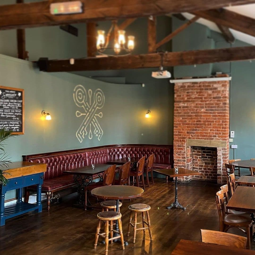 The Cross Keys cozy pub interior fireplace wooden beams Interior of The Cross Keys showing a cozy pub atmosphere with dark wooden tables, red leather bench seating, a brick fireplace, and a unique crossed keys symbol on the wall.