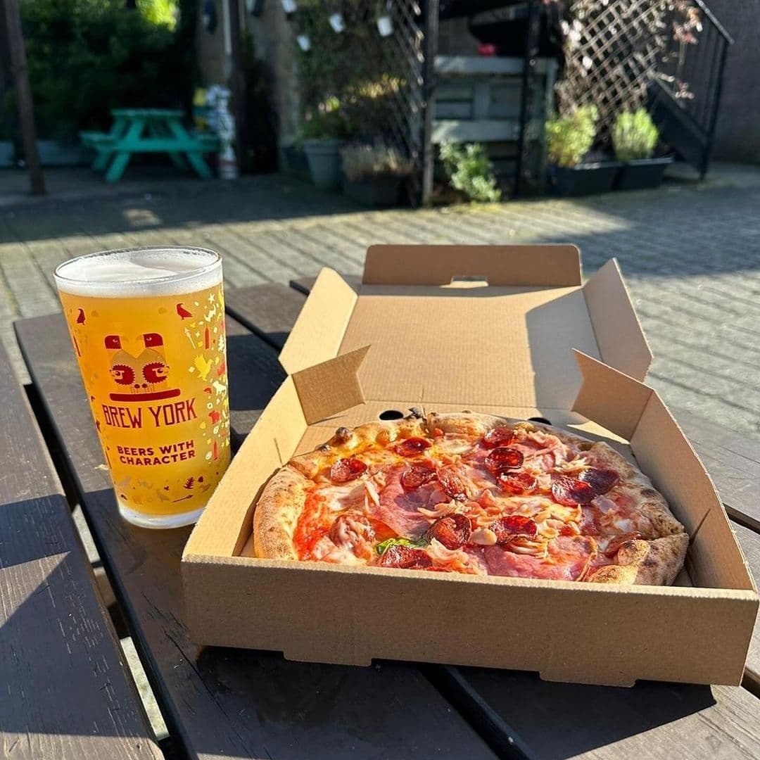 The Horse Farrier Outdoor Dining Pizza Beer Sunshine A fresh pizza in a cardboard box and a pint of beer on a table at The Horse & Farrier outdoor seating area with garden in the background.