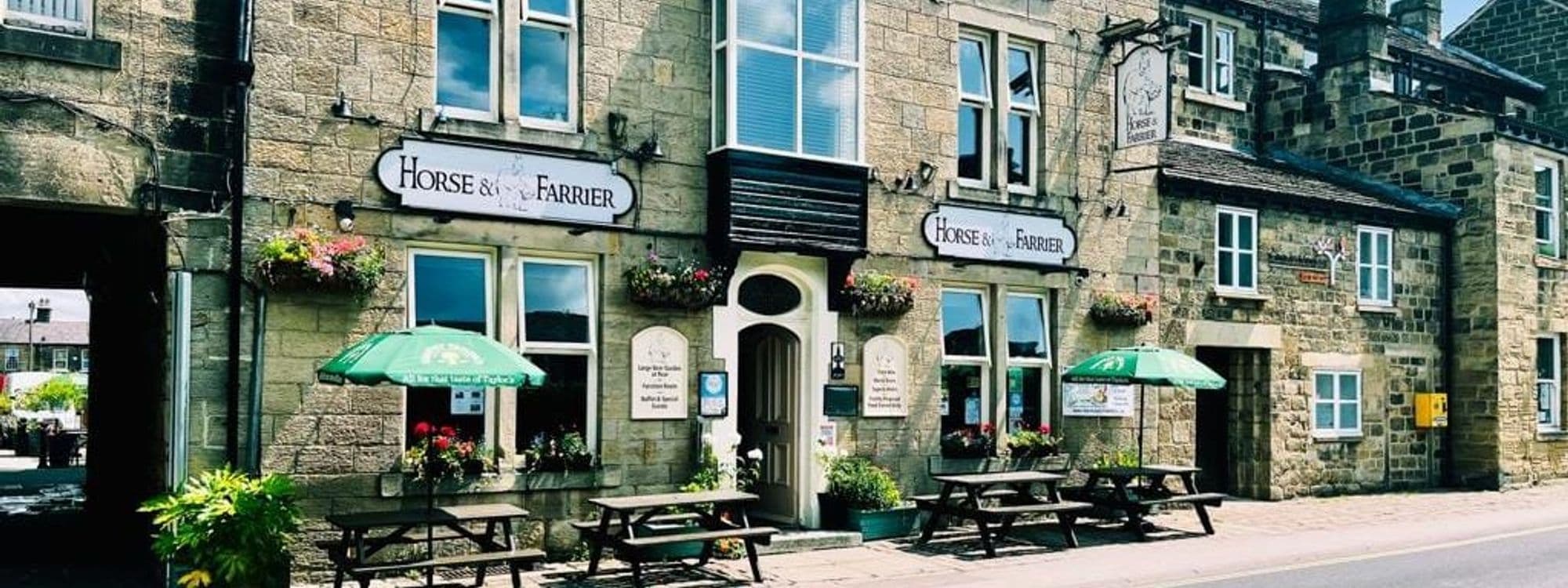 Exterior view of The Horse & Farrier, a traditional stone-built pub with signage, flower baskets, green umbrellas, and wooden benches on the sidewalk.