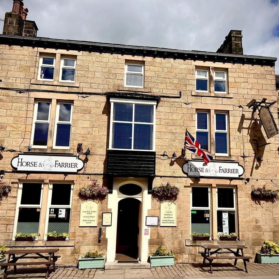 The Horse and Farrier Historic Inn Facade Flag Benches Exterior view of The Horse & Farrier, a traditional stone-built pub with a British flag, hanging sign, and outdoor seating under a clear sky.