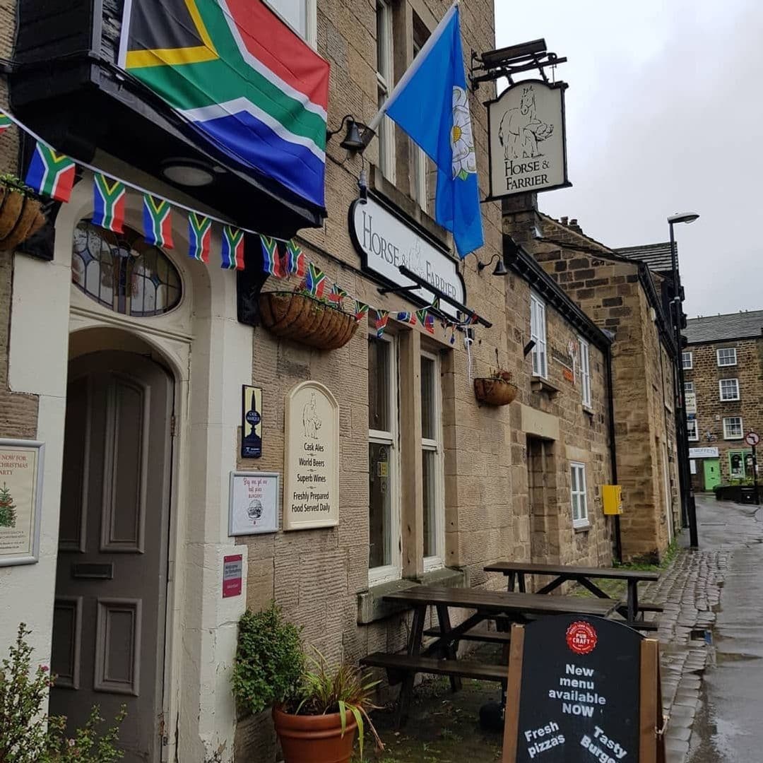 The Horse and Farrier Traditional Pub Exterior Flags Exterior view of The Horse & Farrier, a traditional stone building with a hanging pub sign, adorned with international flags and a blue Yorkshire flag, offering cask ales, world beers, and freshly prepared food.