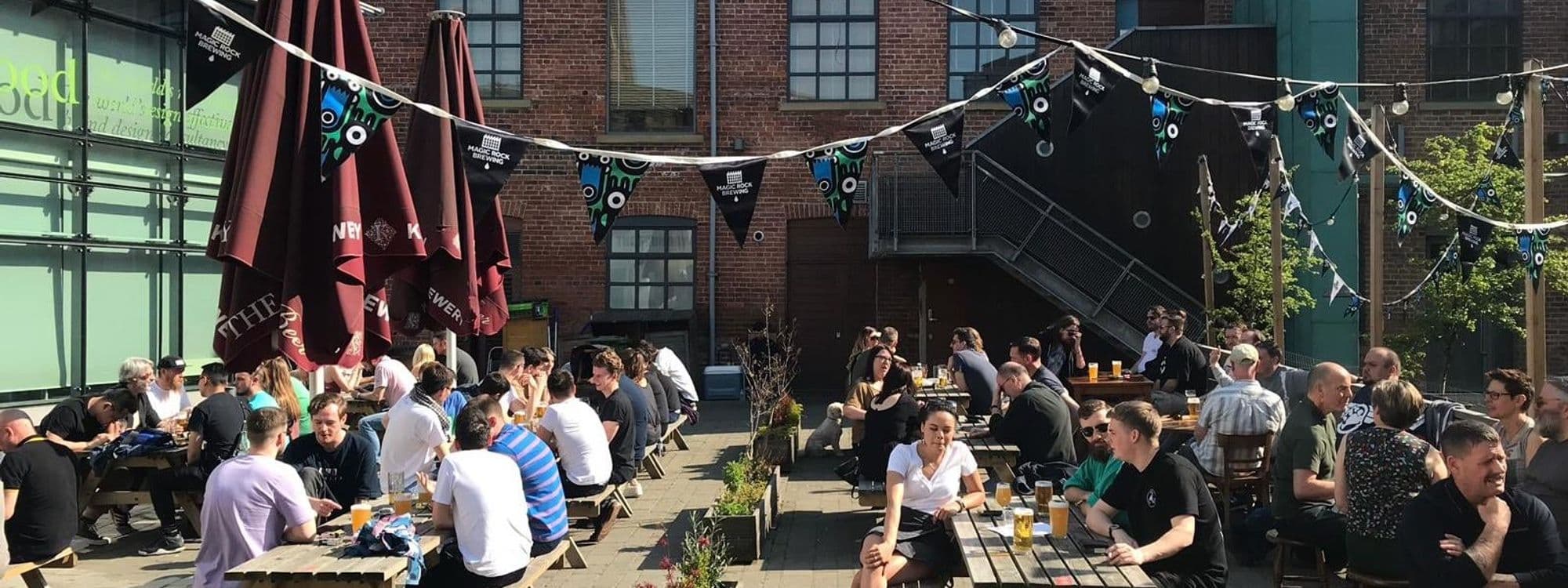 Outdoor seating area at The Cross Keys with people enjoying drinks and conversation under sunny skies, flanked by umbrellas and festooned with string lights and brewery pennants.