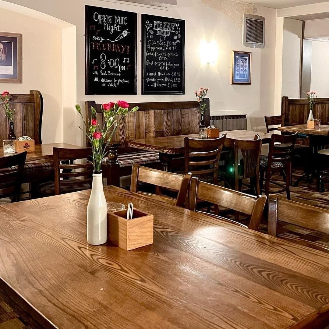 The_Horse_and_Farrier_Cozy_Pub_Interior_Wooden_Tables Interior of The Horse & Farrier showing wooden tables and chairs, a chalkboard advertising events and menu, and vases with red flowers.