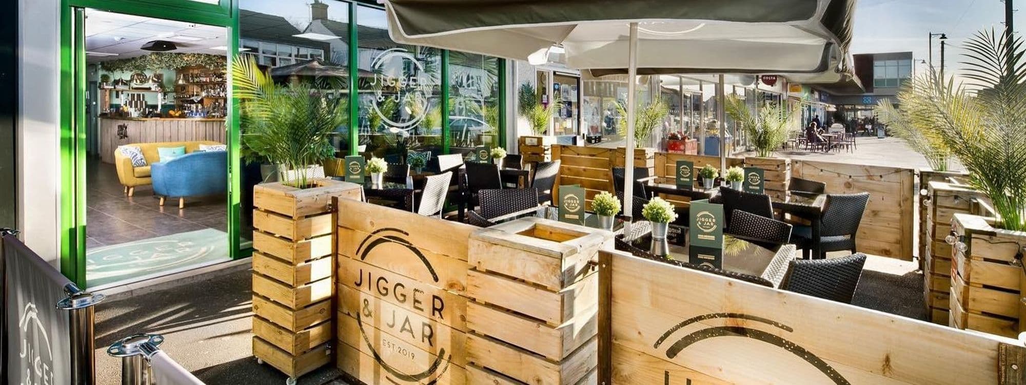 Outdoor seating area of Jigger & Jar with wooden crates, black wicker chairs, and green plants under a sunny sky.