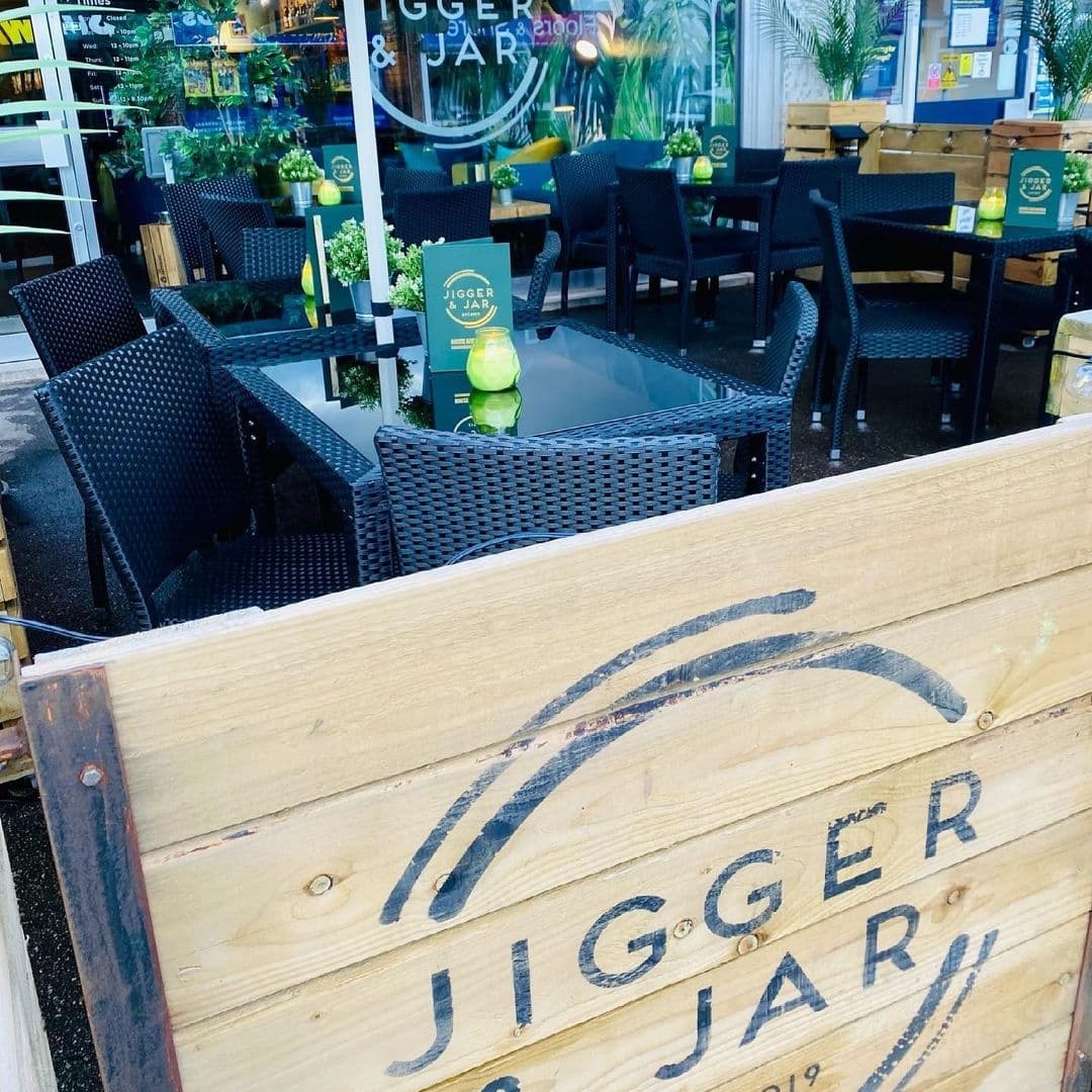Jigger and Jar Outdoor Seating Area Relaxing Atmosphere Outdoor seating area at Jigger & Jar with black wicker chairs and tables, and a wooden barrier featuring the establishment's name.