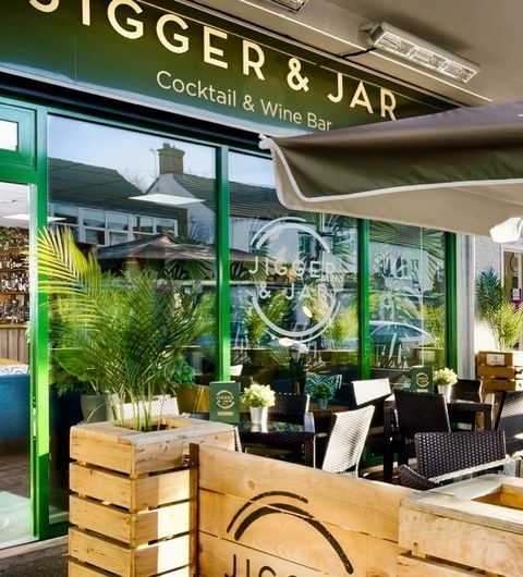 JiggerAndJar CocktailWineBar OutdoorSeating SunnyDay Exterior view of Jigger & Jar Cocktail & Wine Bar with outdoor seating area, wooden crates, and green awnings.