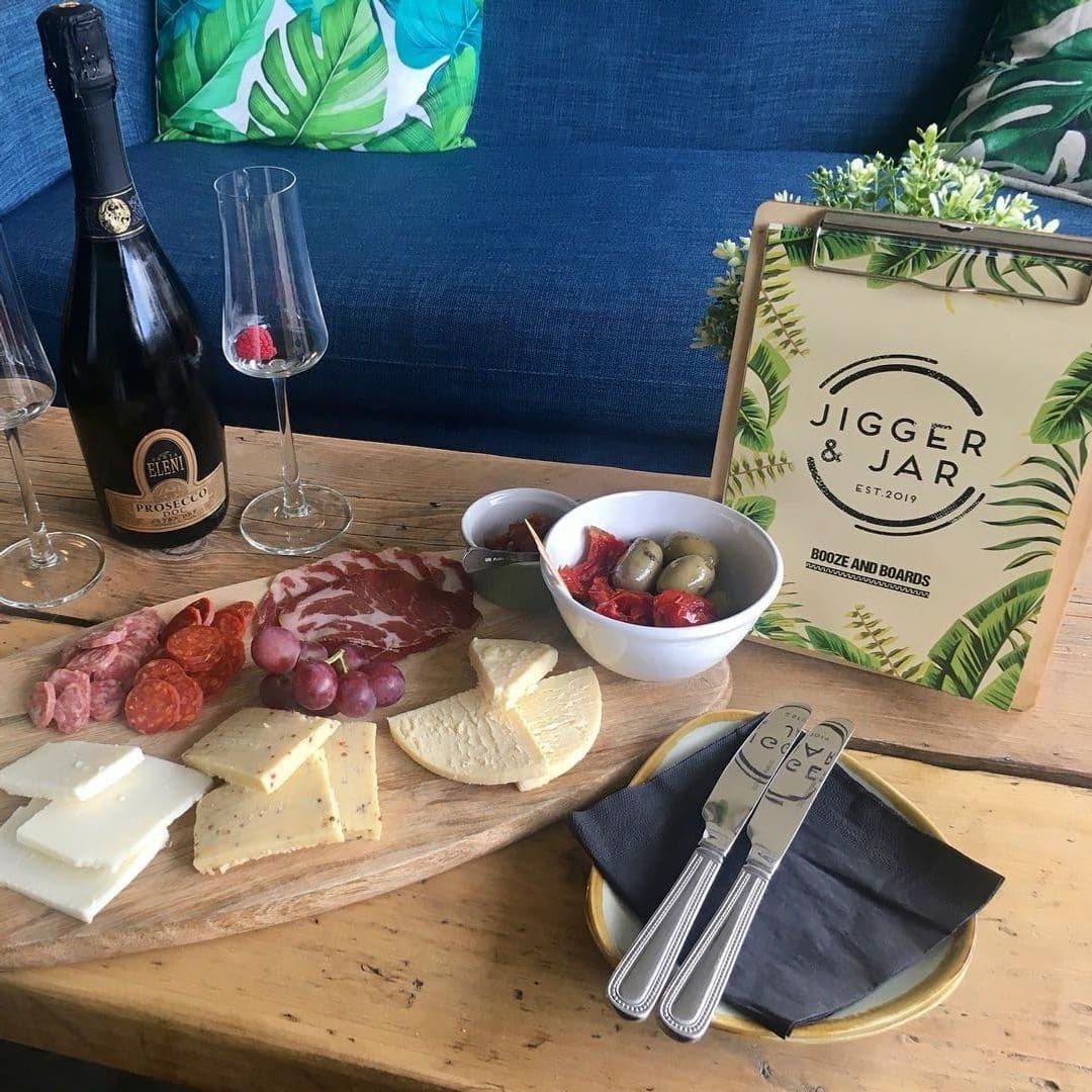 JiggerAndJar_Charcuterie_Cheese_Prosecco_CasualDining Charcuterie and cheese board with a bottle of Prosecco at Jigger & Jar, featuring assorted meats, cheeses, olives, and sun-dried tomatoes on a rustic wooden table with a blue sofa and tropical cushion in the background.