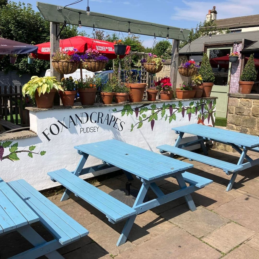 Fox and Grapes Pudsey Sunny Outdoor Seating Floral Decor Sunny outdoor seating area at Fox & Grapes – Pudsey with blue picnic benches, colorful hanging flower baskets, and a white wall with the pub's name and grapevine decorations.